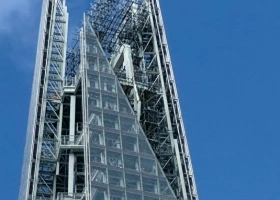 A close-up, low-angle shot of the glass and steel pinnacle of a modern skyscraper, showing the intricate structural framing against a clear blue sky.