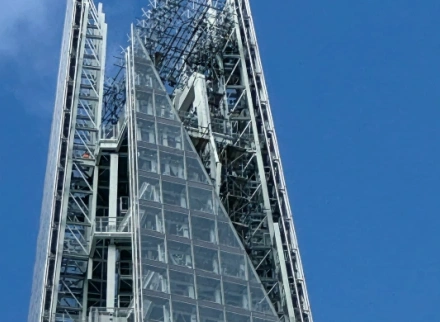 A close-up, low-angle shot of the glass and steel pinnacle of a modern skyscraper, showing the intricate structural framing against a clear blue sky.